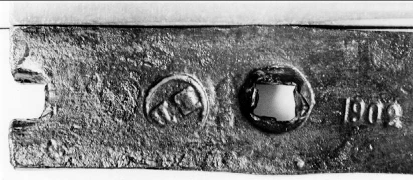 Avhugget stycke av järnband, byggnadsjärn, med ett och ett halvt fyrkantigt hål, stämplat med en rund stämpel EB och 1902..