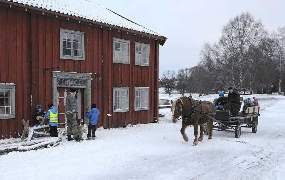 Hest med slede kommer opp på langsida av et rødmalt hus i to etasjer. Snø på bakken og på taket. Utenfor huset står noen små barn og venter på skyss.