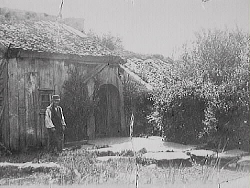 Ryggåsstuga med s k skunke vid entrén. Framför huset är marken belagd med stenhallar och där står en man i väst och keps. Bilden märkt "Carl Heintz".
