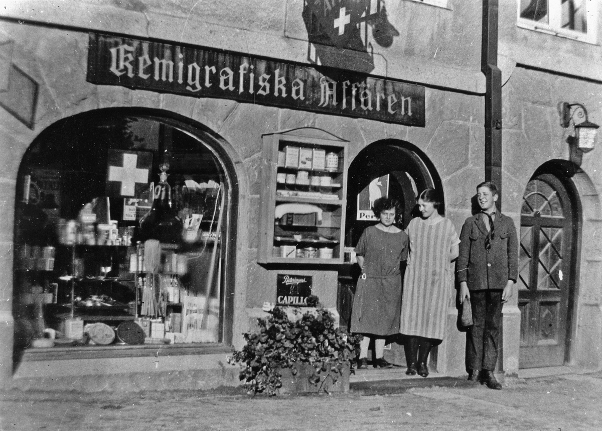 Signhild Skog, Gunhild Svantesson och Carl Ivar Landgren (här ca 15år) står utanför Kemigrafiska Affären på Göteborgsvägen. Kvarteret Cederberg 3. Knut Landgrens Kemikalieaffär, blev sedemera Landgrens Färg och Parfymeri, 1922-1984-04-01. Efter det låg Bäcks Hår och Parfym, i samma lokaler, Magasinsgatan 1.