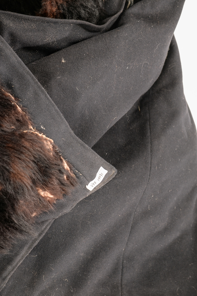 Frakk, kjørepels i svart ulltøy, fóra inni med svart bjørneskinn, ermane er også fòra med bjørneskinn. Ulveskinn(?) på kragen, slaga og rundt ermane.  Tre knappar og hempe på kvar side i opninga. Ein knapp til å halda saman kragen med.