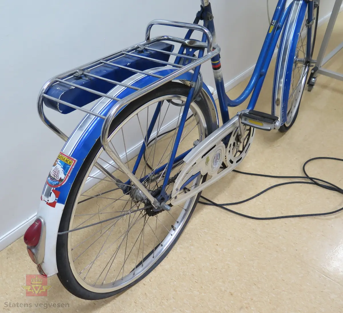 Sykkel, damemodell. Sykkelen er blå og hvit med brunt sete og hvite styreholker. Sykkelen er utstyrt med bagasjebrett, ringeklokke, frontlykt og refleks bak. Sykkelen har lås med nøkkel.