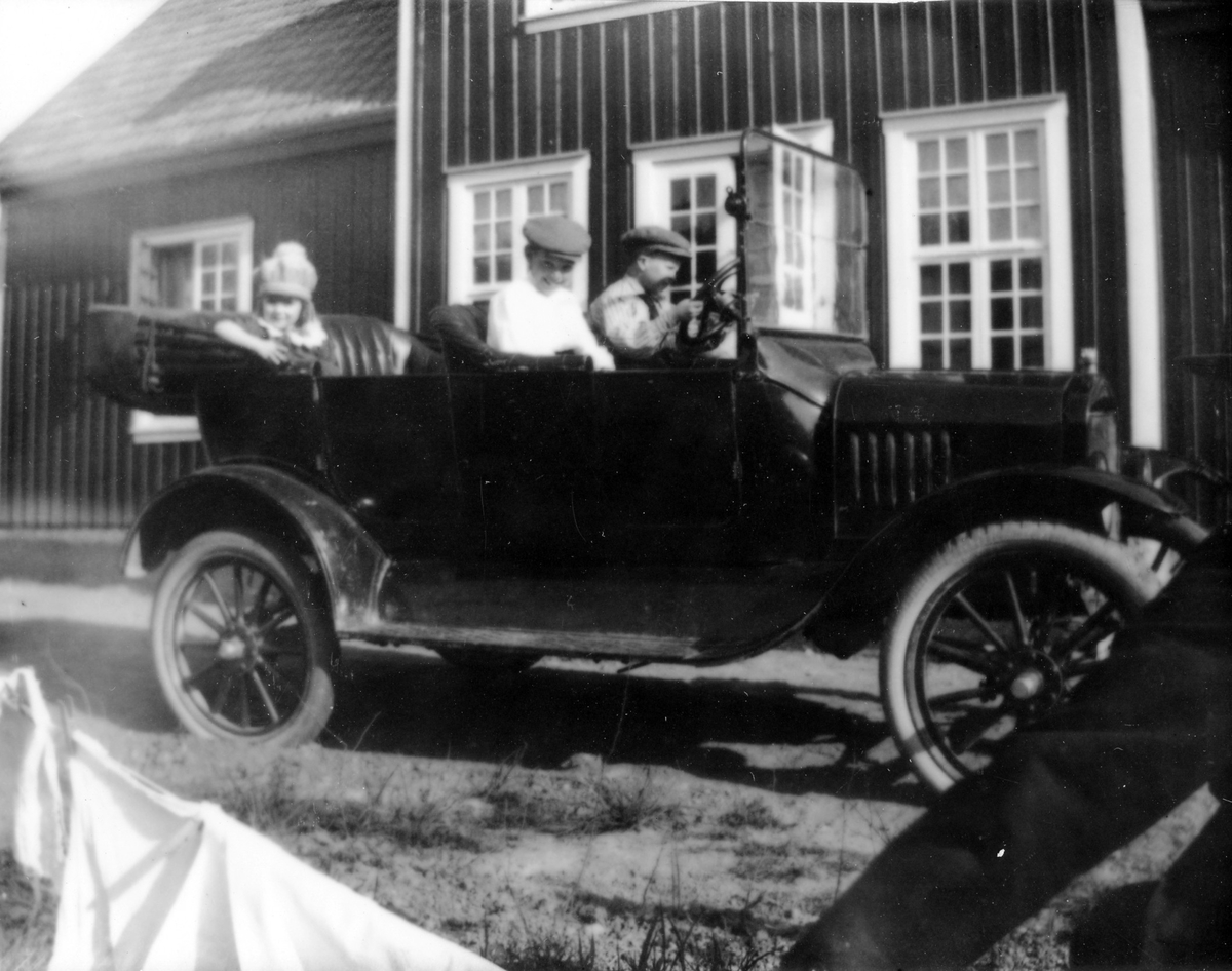 En bil med sufletten nerfälld står parkerad framför ett hus. I bilens framsäte sitter två pojkar och i baksätet en flicka.