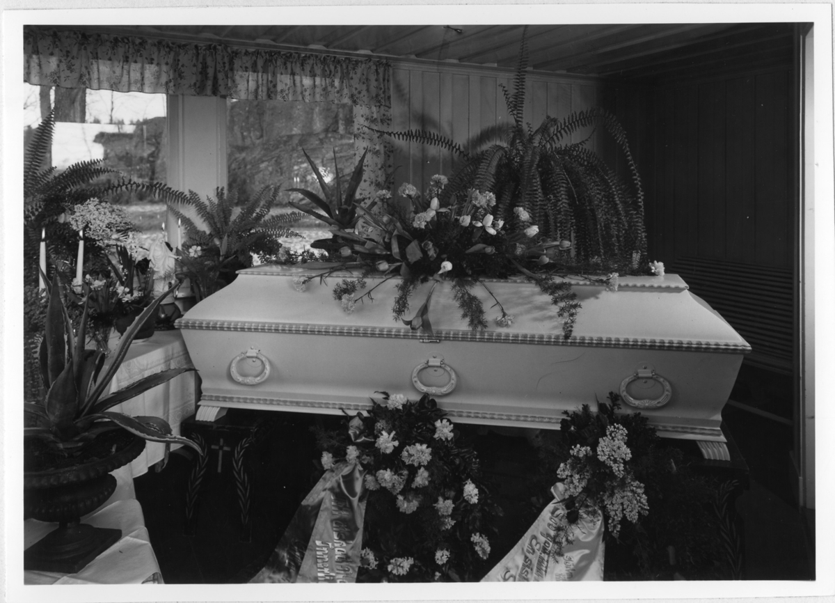 En kista täckt med begravningsblommor, en liten statyett av Jesus står i fönsterkarmen.

Begravning år 1947 i Alingsås.