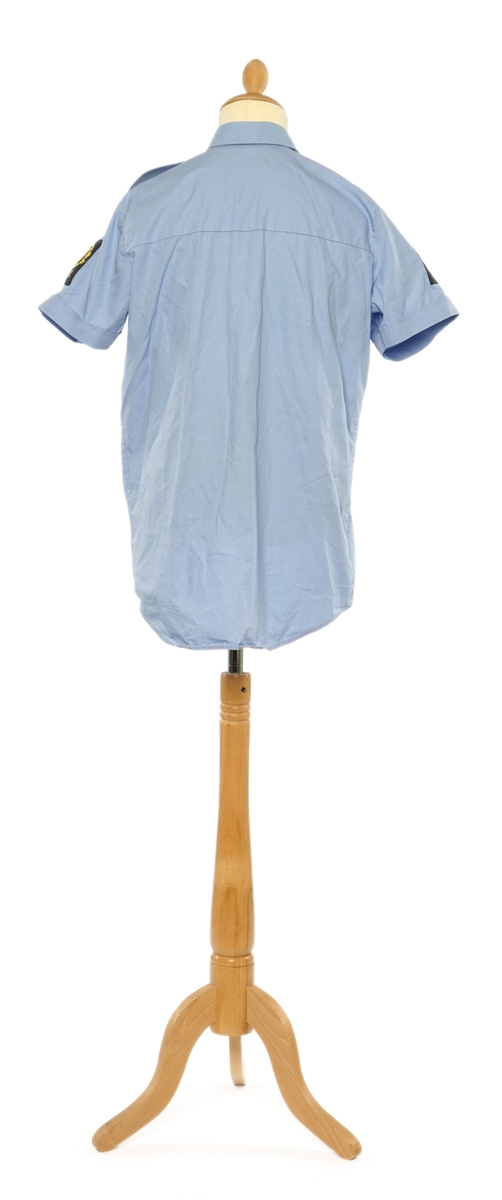 Lys blå  kortermet skjorte av et fintvevd stoff. Epauletter uten tilsatt distinktsjon. Tekstil merke på begge armer: riksløve med hvit øks omgitt av eikeblader over TOLL. Tekstil merke på venstre bryst, gul bokstaver på sort, 'TOLL'.
