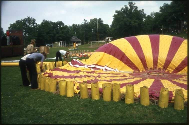 75-årsminnet av Andrées Nordpolsfärd firas med Ballongdagen på Åsavallen i Gränna 1972. En ballong ligger utlagd på gräsplan med gula ballastsäckar runtom, den håller på att fyllas. Ballongen är möjligen gasballongen Gatzweiler Alt. I bakgrunden skymtar dansbanan. Diabild.