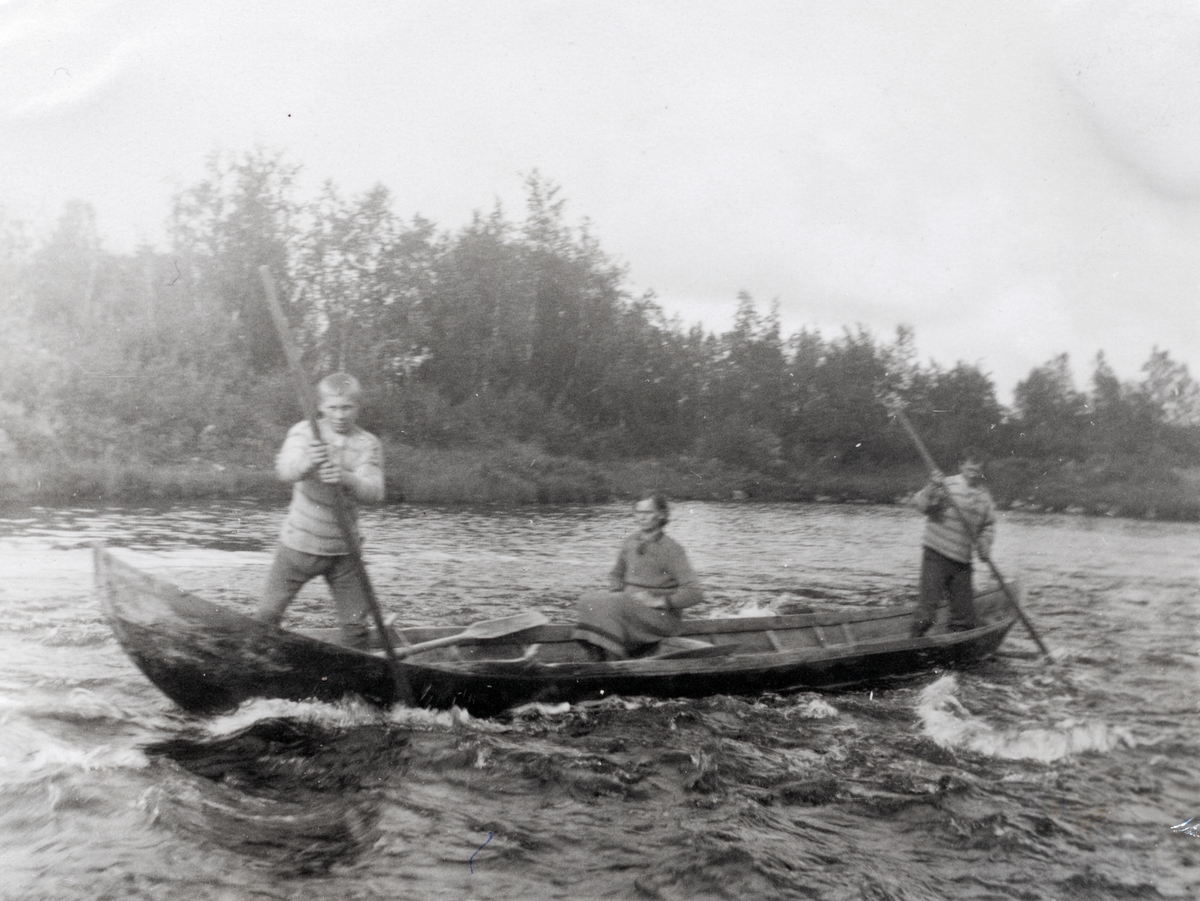 Motiv av to menn og ei kvinne i en elvebåt i Kautokeinoelva. Mennene staker mot stømmen i elva.