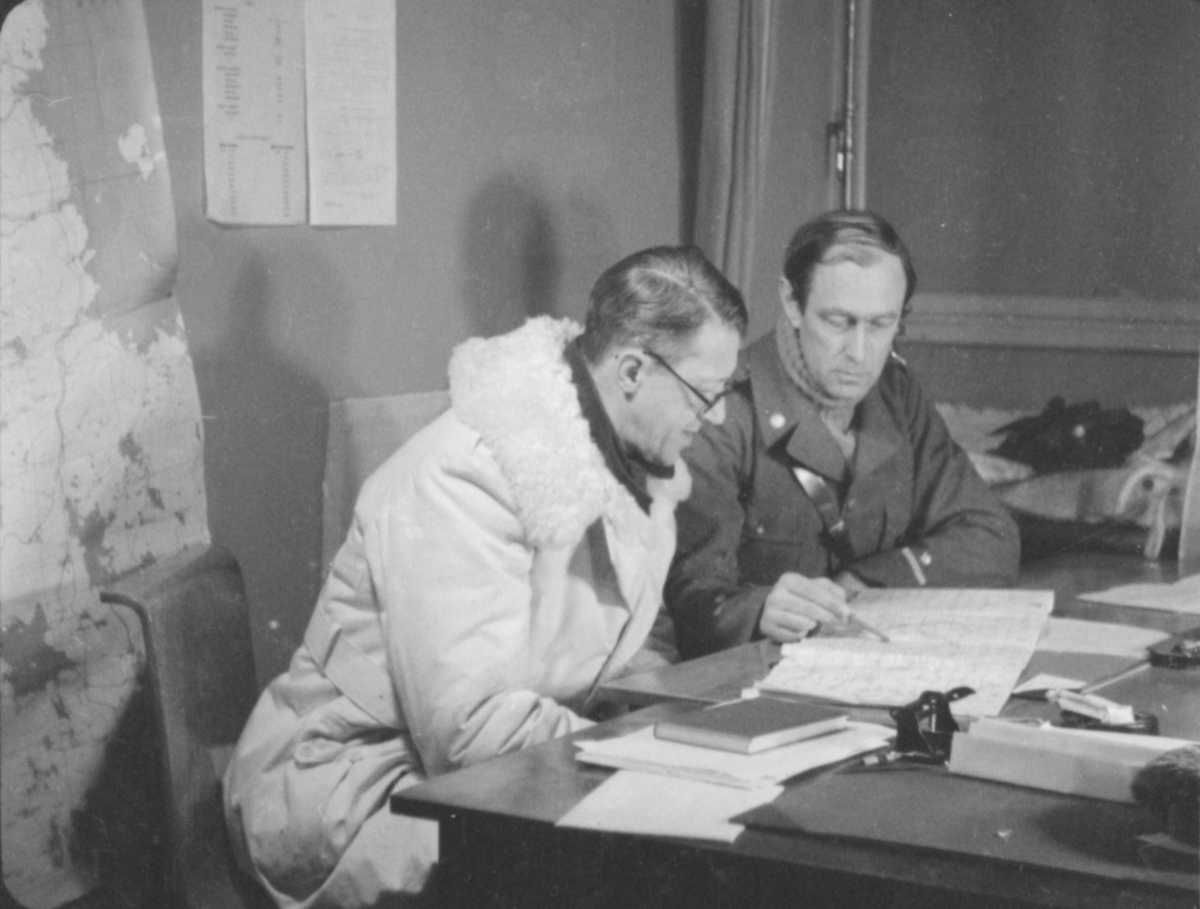 Flottiljchef Hugo Beckhammar (till höger) visar en militär i vinteruniform en lista, sittandes vid ett skrivbord. Vid Svenska frivilligkåren i Finland, F 19.