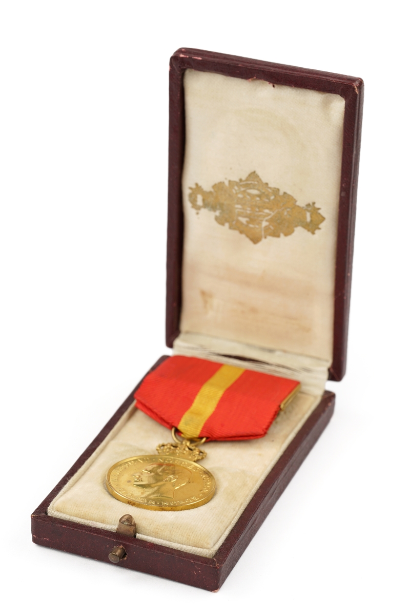 Gullmedalje med krone festet i et tekstilbånd med festenål liggende i etui