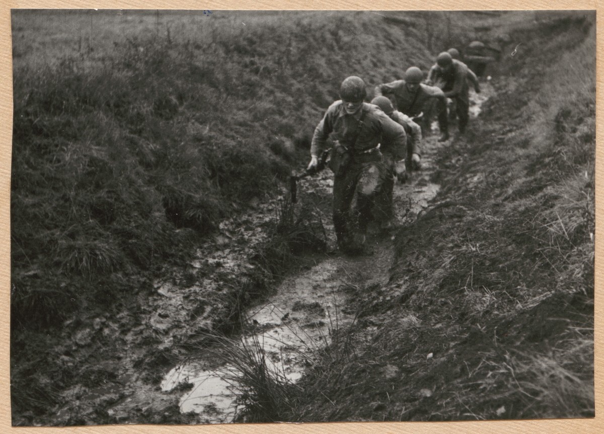 Bilden visar en grupp värnpliktiga som springer i en lerig dike.