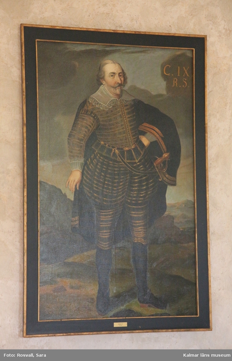 Karl IX i helfigur, stående, i mörk dräkt med guldstickningar, mantel med guldbroderier, spetskrage samt spetsar vid handlederna. Värja, svarta strumpor och skor med spännen.