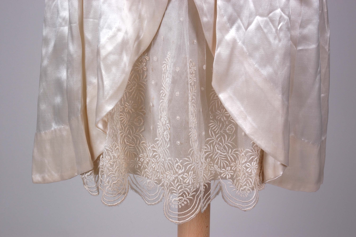 Knelang brudekjole i blank, benhvit silkesateng. Dekorert med innfellinger av tyllblonde.