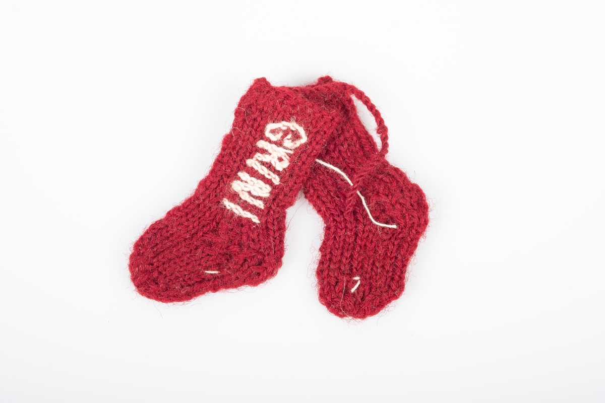 Ett par strikket sokker som er bundet eller sydd sammen. Sokkene er strikket med garn i burgunder. Det er brodert "Grini" på den ene sokken og "1943" på den andre, med hvitt garn.