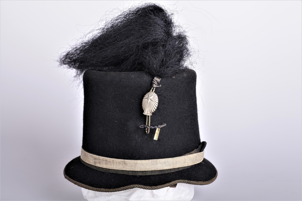 Militærhatt, svart hårhatt, silkebord, hårdusk framme, silkefór. Brukt av pensjonerte underoffiserer fra ca 1870-årene og utover.