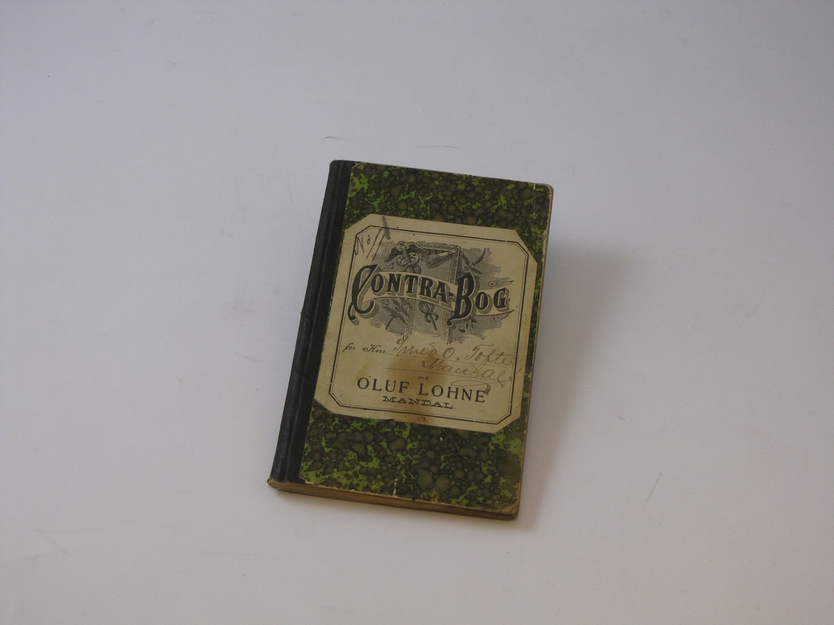 Bok med grønt og lysegrønt papiromslag med flekkete mønster, boken har også svart tektstil ryggbinding. Det er en lapp klistret på framsiden skrift.
