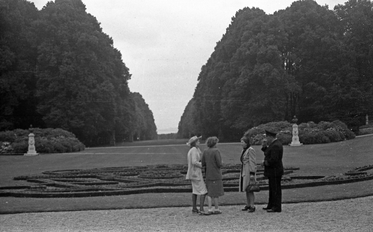 En mann i uniform og tre kvinner i Fredenborg slotts park. Store grønntområder med høye trær på begge sider. Suderøy på vei til fangstfeltet.