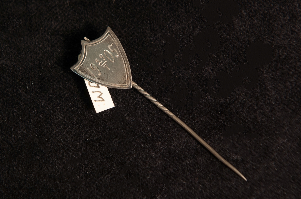 Sköldformad kråsnål av metall (silver ?) med graverad datering: "19 29/7 05". Ostämplad.