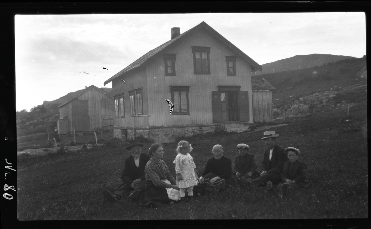 Portrett av familie sittende på gårdstun foran hus.