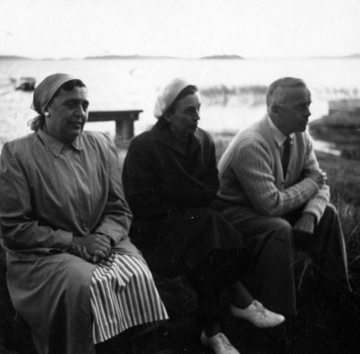 Årsmöte vid en strandkant där två kvinnor och en man sitter på en bänk. Anteckning på kortets baksida "Årsmöte 1954".