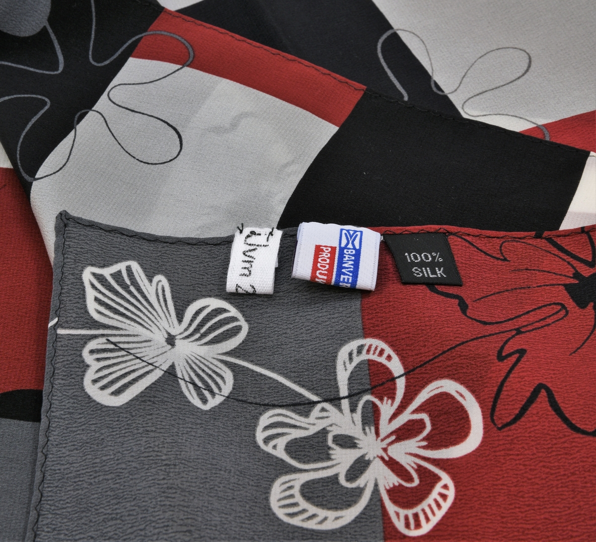 Scarf av silke med ett upprepande mönster bestående av blommor och geometriska figurer i rött, grått, vitt och svart. Scarfen har två lappar fastsydda vid ena kortsidan.