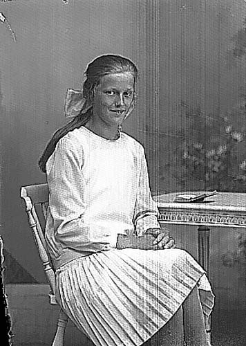 Elna Magnusson i ljus klänning/plisserad kjol sitter vid ett bord. Sannolikt i samband med konfirmation.