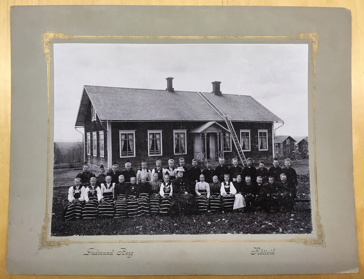 Folkskolelärarinnan Charlotta Kristina Johansson med skolbarn, delvis i folkdräkt, sittande framför skolhuset. Troligen vid Röjeråsen i Rättvik.
