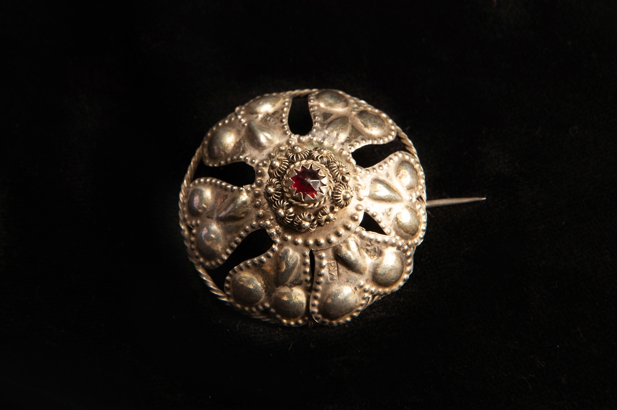 En rund malja av ciselerat silver med utseendet av hjärtformade blomblad kringen en infattad röd sten av glas. Runt stenen filigranarbete. Spännet, maljan, har gjorts om till en brosch.