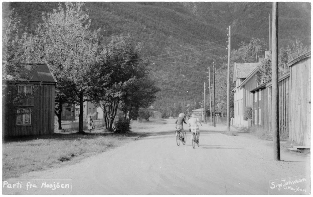 Postkort med parti fra Mosjøen. Nedover Elvegata. Vi kan skimte Fru Haugans Hotell i enden av gata til høyre. To personer på sykkel.