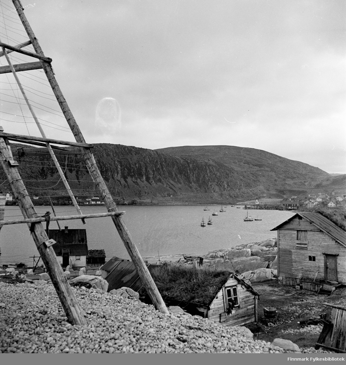 Kjøllefjord 1940. I forkant står det en fiskehjell, bildet viser utsikten  innover mot Kjøllefjord med et torvhus i forgrunnen, bakover sees trehusbebyggelse og fiskebåter oppankret i Kjøllefjorden.