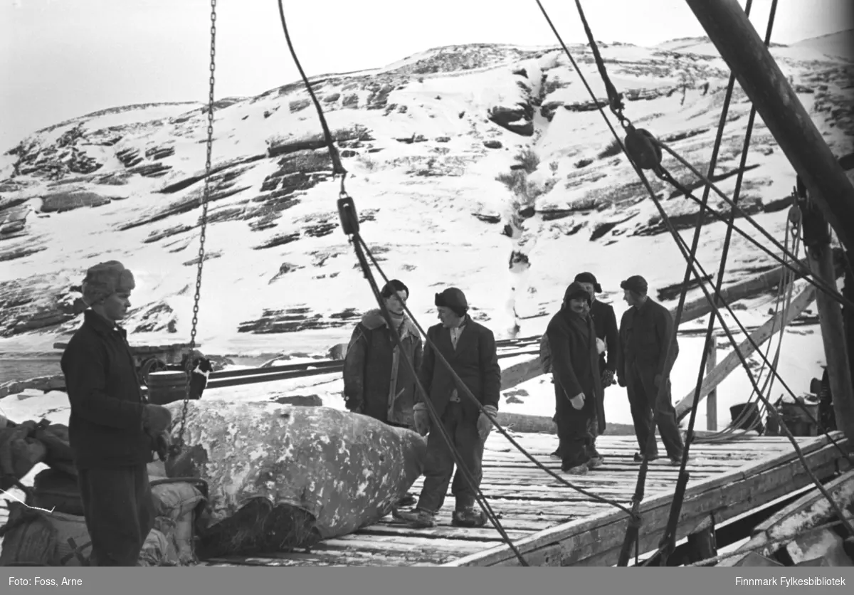 Arkitekt Arne Foss har fotografert dette bildet fra båten ved Reinelva, Repvåg i februar-mars i 1947. Seks menn står på kaia. Personen nr 2 fra venstre er  Ivar Utsi, nr 3 foran er Samuel Josefsen, Stranda. Gjenreisning er i gang i Finnmark. Varer er lastet på en kai, bl.a. en stor høylass. 