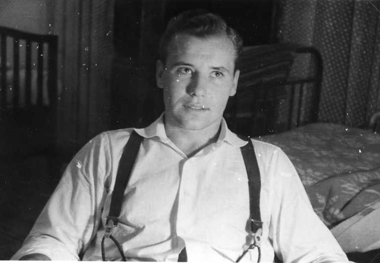 Porträtt av en ung man i vit skjorta och hängslen. Sitter in en sängkammare, en säng i bakgrunden.