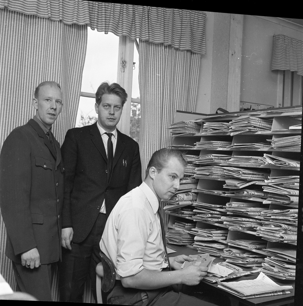 Premiär för lantbrevbärare i Gränna, 1 okt 1967. Henry Kvist och Rolf Lindén stående bakom Anders Hjelte som sitter i vit skjorta och slips vid ett bord med post i händerna. På bordet en hög hylla med postfack fyllda med tidningar.