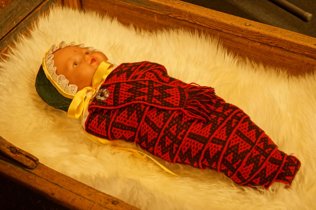 Dukke reivet med et reivebånd i svart og rødt. Dukken har også på seg et rekontruert hodeplagg og ligger i en vogge.