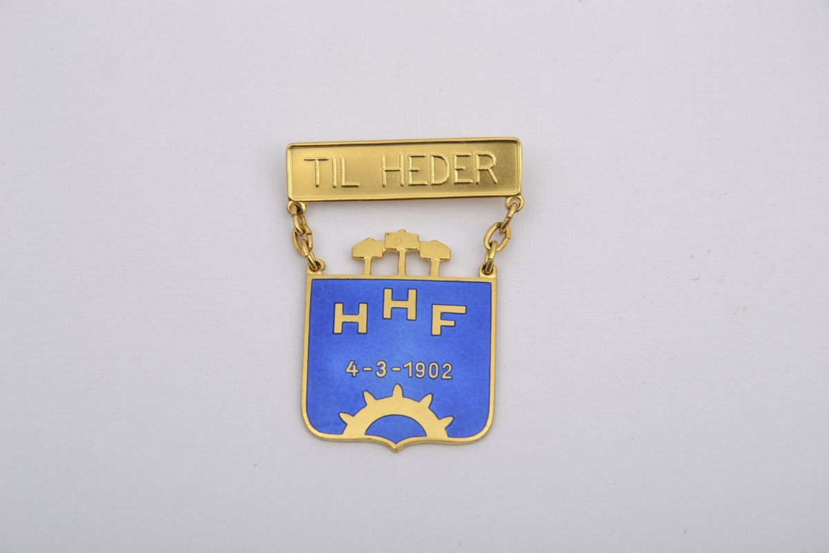 Medalje for hedersbevisning med Harstad Håndverkerforenings sin logo framstilt i gullbelagt sterlingsølv og blå emalje med datoen 4-3-1902 og en nål hvor det står TIL HEDER. Ligger i originaleske.