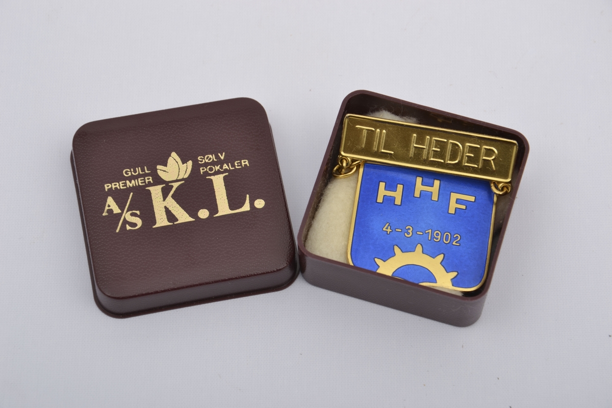 Medalje for hedersbevisning med Harstad Håndverkerforenings sin logo framstilt i gullbelagt sterlingsølv og blå emalje med datoen 4-3-1902 og en nål hvor det står TIL HEDER. Ligger i originaleske.