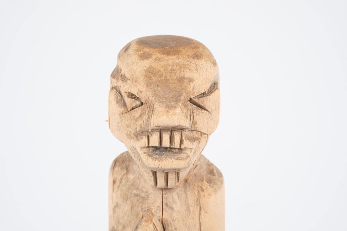 Figur skåret ut av en stubbe. Figuren kan minne om skulpturer fra Søramerikanske stammer. Figuren er av et menneske som sitter på en stubbe med håndlfatene slått sammen.