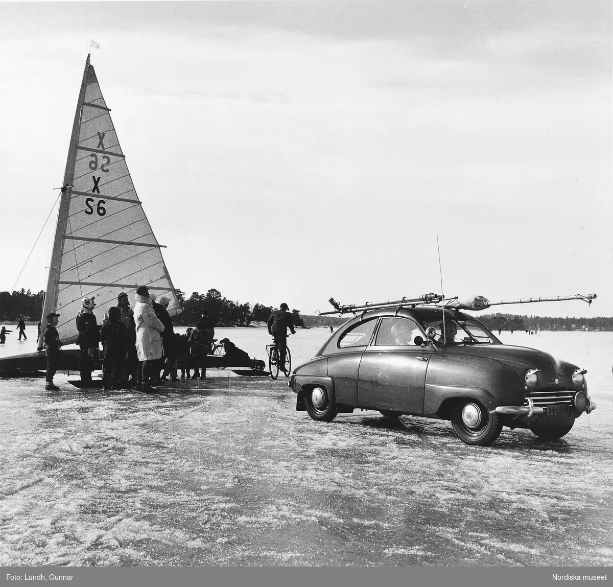 Människor står kring en isjakt på en frusen vik på Mälaren år 1954. En Saab 92 står på isen.