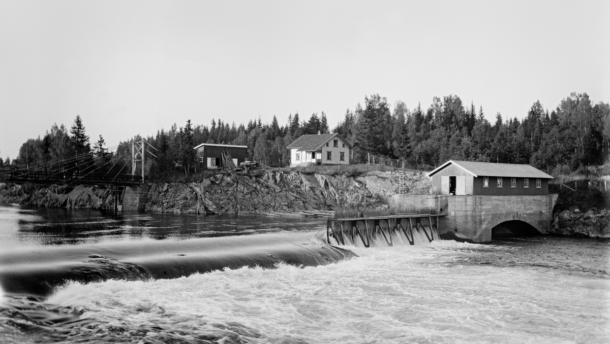 Bygging av nåledam - såkalt «fermettedam» - ved Svanfossen i elva Vorma i Fenstad i Nes kommune på Øvre Romerike, fotografert i 1916.  Den øvre delen av dette vassdraget ble noe regulert i slutten av 1850-åra, for å gjøre det mulig å kople den da nyetablerte hovedbanen med dampskipsruter på Mjøsa.  Den første dammen ved Svanfossen skal ha vært bygd i 1910.  Her ser vi (til venstre i forgrunnen) hvordan elva faller over en terskel på elvebotnen.  På den østre delen av denne var det montert en såkalt «fermettedam», en stålkonstruksjon som bærer ei gangbru, der fløterne kunne gå når de skulle «sette dammen» (stenge) eller «sprette» (åpne) den. Dette ble gjort ved hjelp av «nåler», plank som ble stukket mot strømmen, slik at den lå an mot en terskel i botnen av elveløpet og mot gangbrua.  Disse nålene ble satt tett i tett, slik at de sammen dannet en vegg mot vannet.  Når dammen ble åpnet, brukte karene små kjetting- eller taustubber med ei jernnål i enden, som ble tredd gjennom hull i den øvre delen av nålene, og trakk til.  Ved siden av damkonstruksjonen ser vi et rektangulært hus med saltak på en høy mur, med en hvelvet åpning mot elveløpet under.  Dette kan muligens ha vært et lite kraftverk.  På bakkekammen sentralt i bildet lå det et  halvannenetasjes våningshus med kvite ytterveger, og ved siden av dette en rødmalt uthusbygning med saltak.