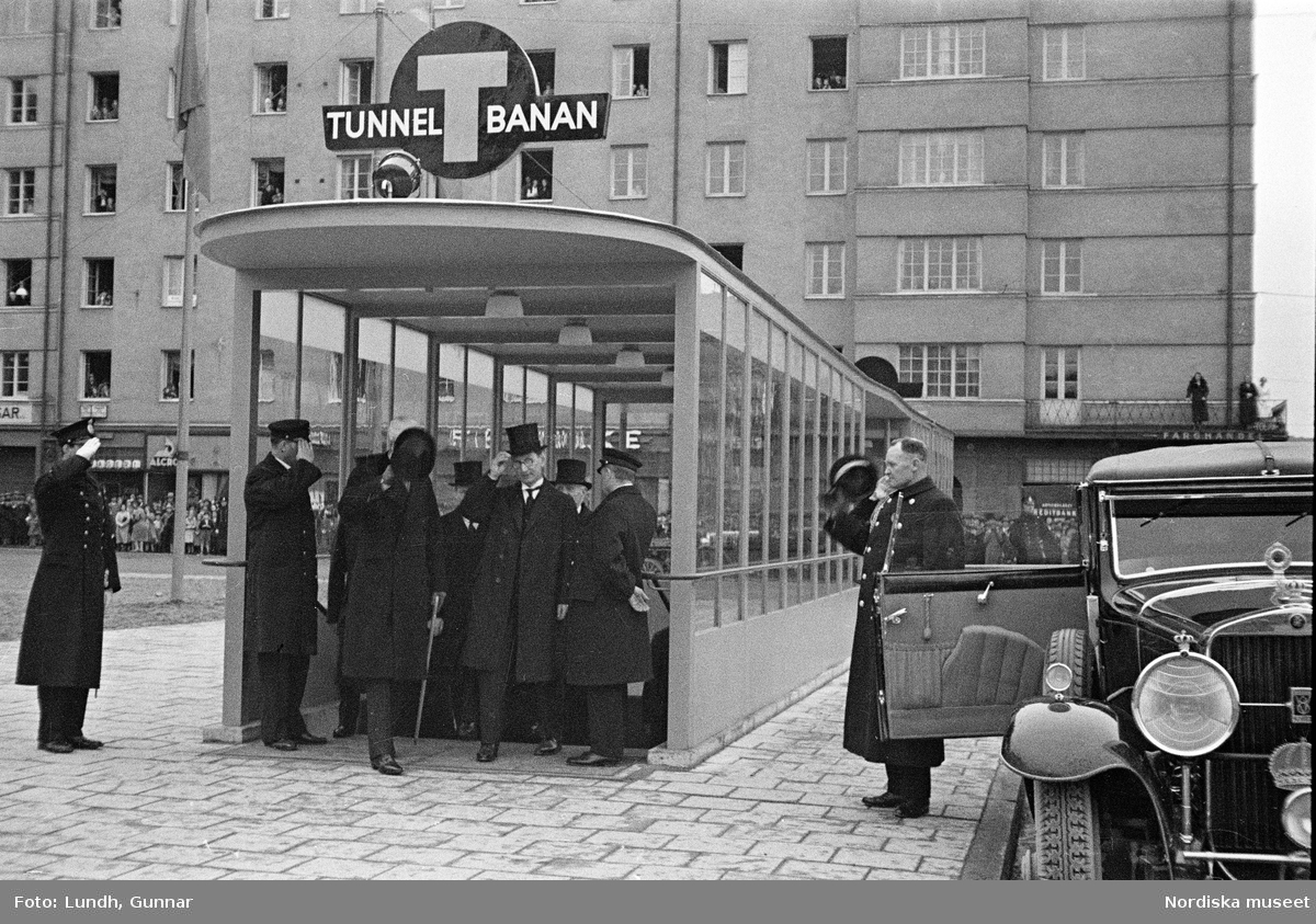 Invigning av Södra Bantorgets (Medborgarplatsen) tunnelbanestation på Södertunneln den 30 september år 1933, Kung Gustaf V kliver in i en bil, porträtt av en man i uniform - en chaufför vid Kungliga Hovstallet, detalj av en bil med lykta och skärm, en grupp män går ut ur en tunnelbanenedgång på Södra Bantorget - män i uniform gör honnör och i bakgrunden står människor i öppna fönster.