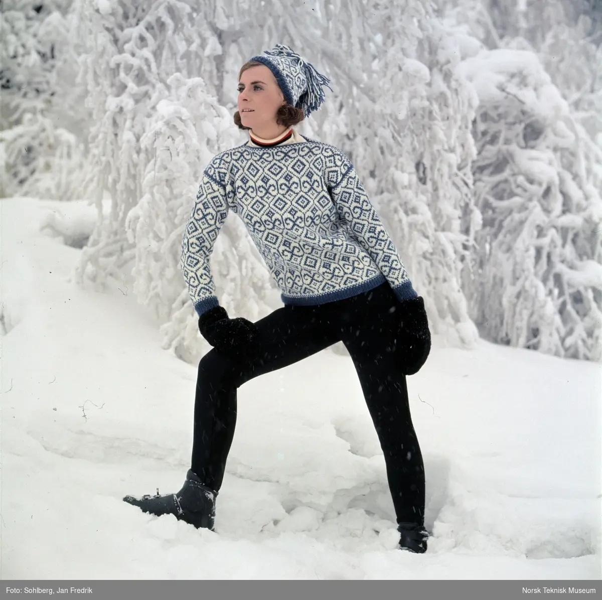 Kvinnelig modell viser vintermote; en blå og hvit-mønstret strikkegenser med tilhørende lue. Fotografert utendørs i vinterlandskap med snø.