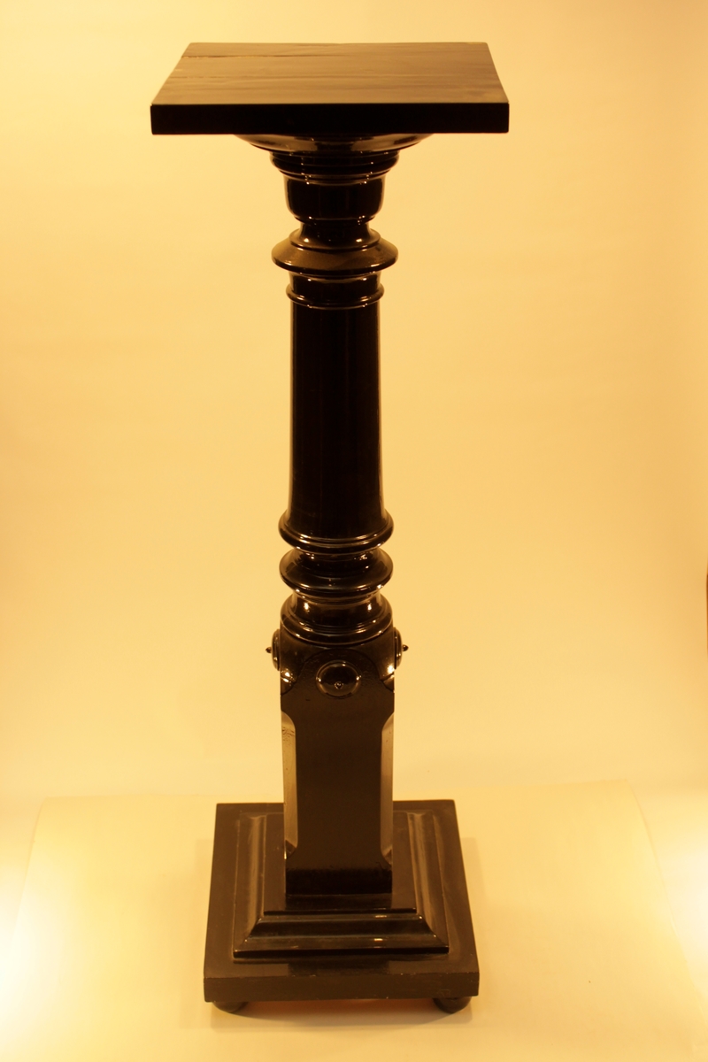Svartmålad helblank piedestal i form av en svarvad stolpe samt kvadratisk skiva för blomkrukan.