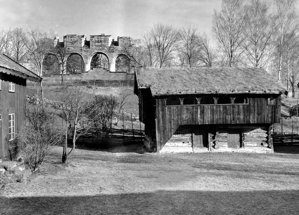 Svart-hvitt foto av høyloftet fra Skråstad gard i Vang etter at huset ble flyttet og gjenreist på museet, domkirkeruinen i bakgrunnen.