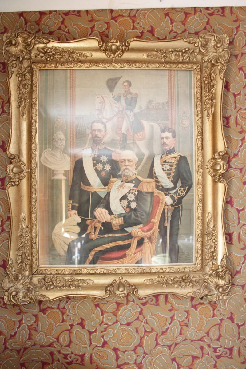 Tryck bakom glas och rikt utsirad förgylld ram. Föreställande kung Oscar II, kronprins Gustav (den V) och prins Gustav Adolf (den VI). I bakgrunden syns Karl XIV Johan till häst på en tavla samt en gipsbyst av kung Gustav II Adolf.