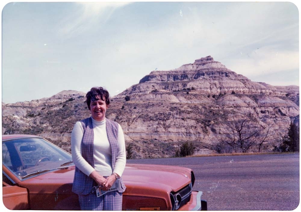 Kvinne står ved bil med fjell i bakgrunnen. "Pat - 1976"