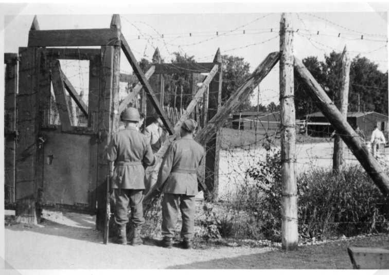 Okänt interneringsläger. På hitsidan står två svenska militärer och bakom högt taggtrådsstängsel syns män i civil klädsel.