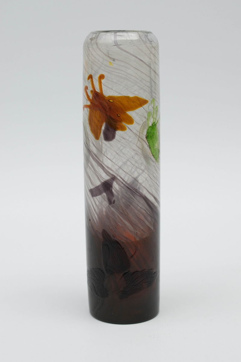 Sylinderformet vase i overfangsglass. Øvre del er i klart glass med innlagt grålilla tråddekor samt marqueterie de verre i form av insekter i brunt og grønt. Den nedre delen er tilnærmet opak, og dekorert med grålilla blad- og plantemotiver på brunfarget bunn.