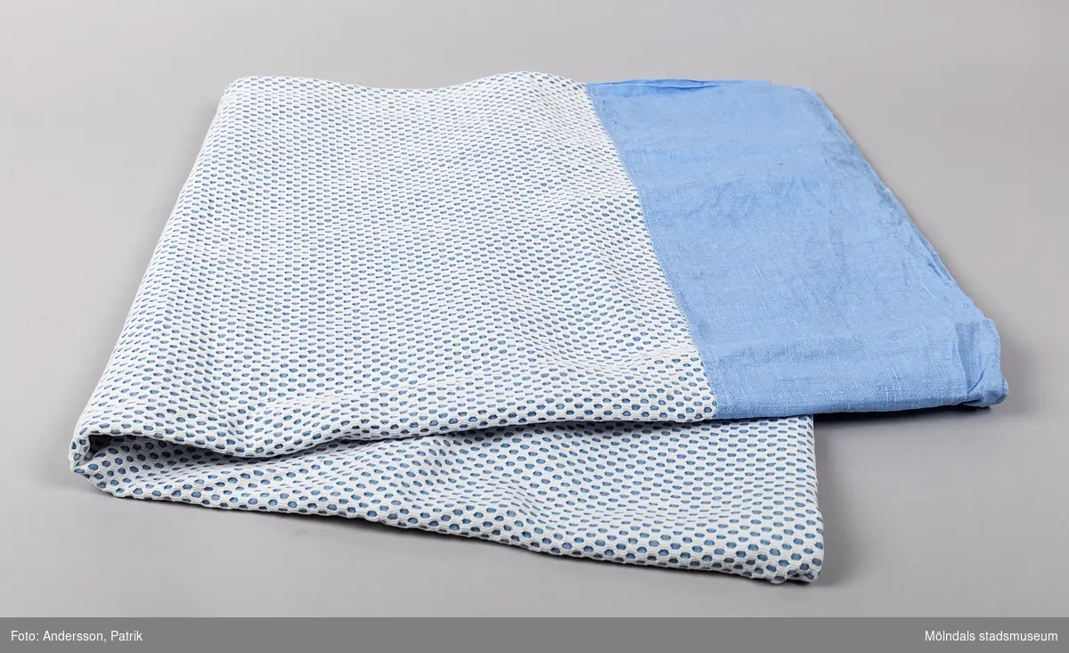 Överkast eller tunnt täcke som består av två lager. Det understa lagret består av en blå lakansväv. Det övre av ett vitt genombrutet tyg.
