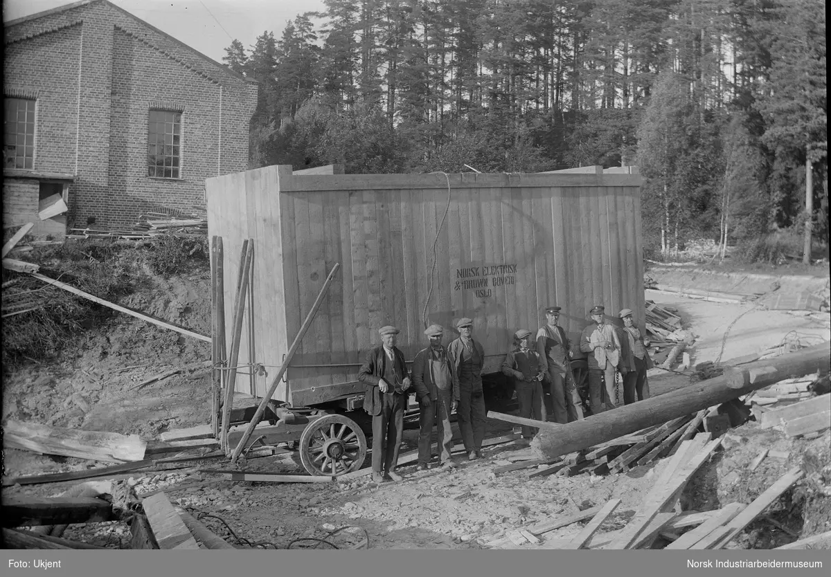 Anleggsarbeid under bygging av Labro Kraftstasjon. Menn oppstilt foran transportvogn til hest. Vognen har påskriften "Norsk Elektrisk & Brown Boveri, Oslo".