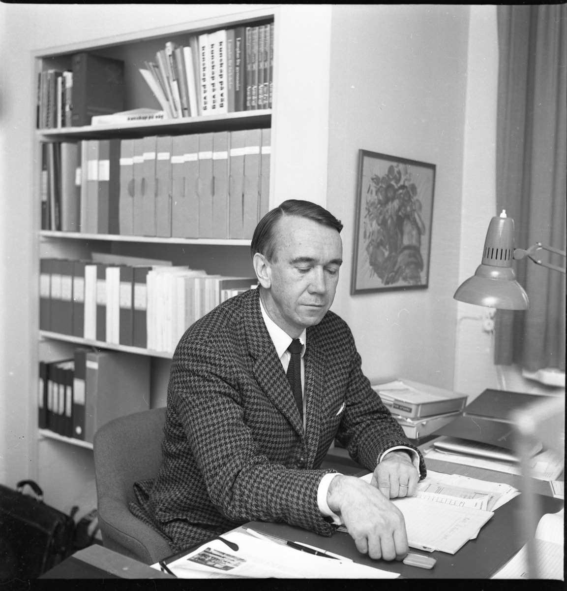 Per-Anders Holkers i rutig blazer och slips sitter vid ett skrivbord med papper på bordet. I bakgrunden en bokhylla med böcker och pärmar.