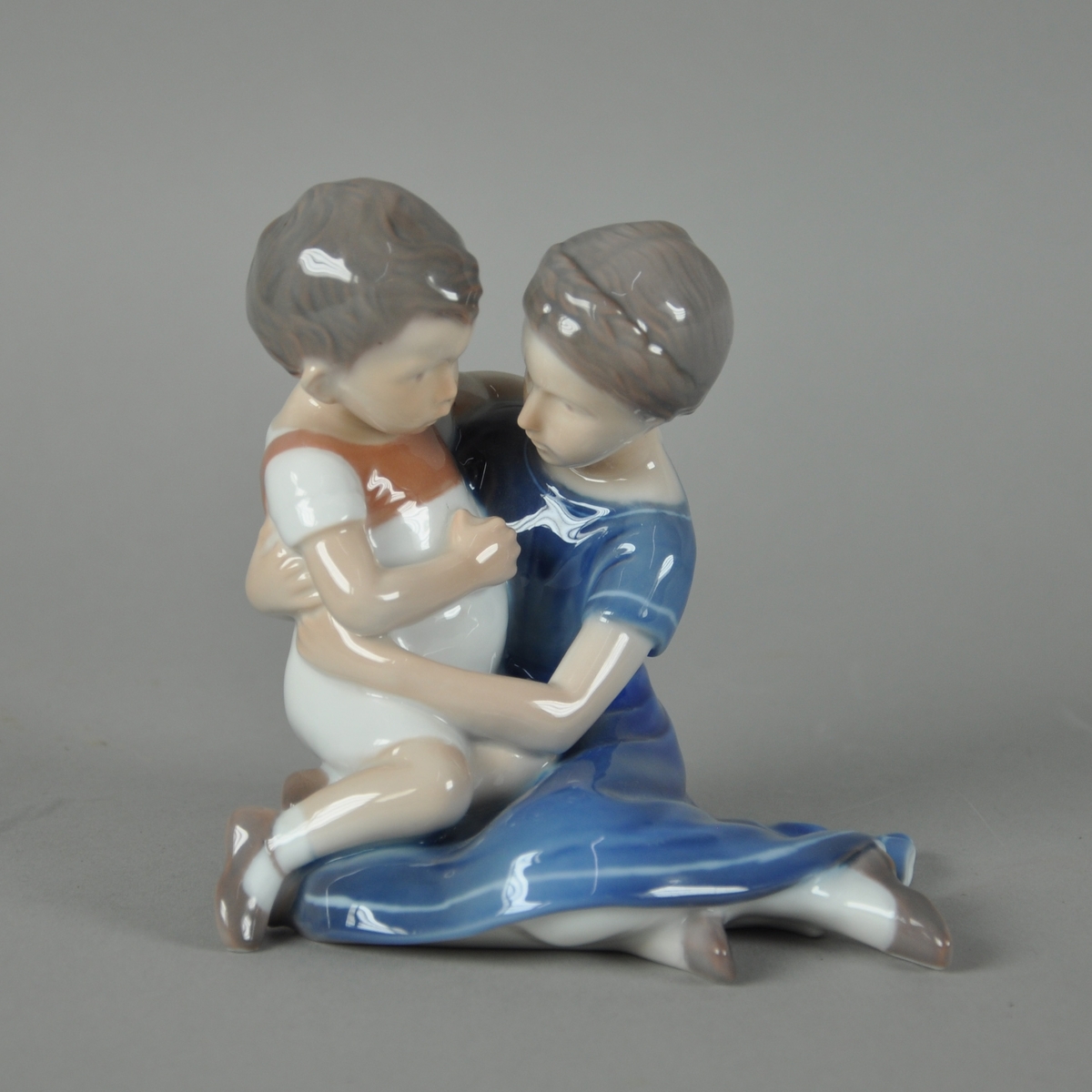 Skulptur av glassert keramikk, med fargedekor. Skulpturen forestiller jente i blå kjole med ung gutt på fanget.